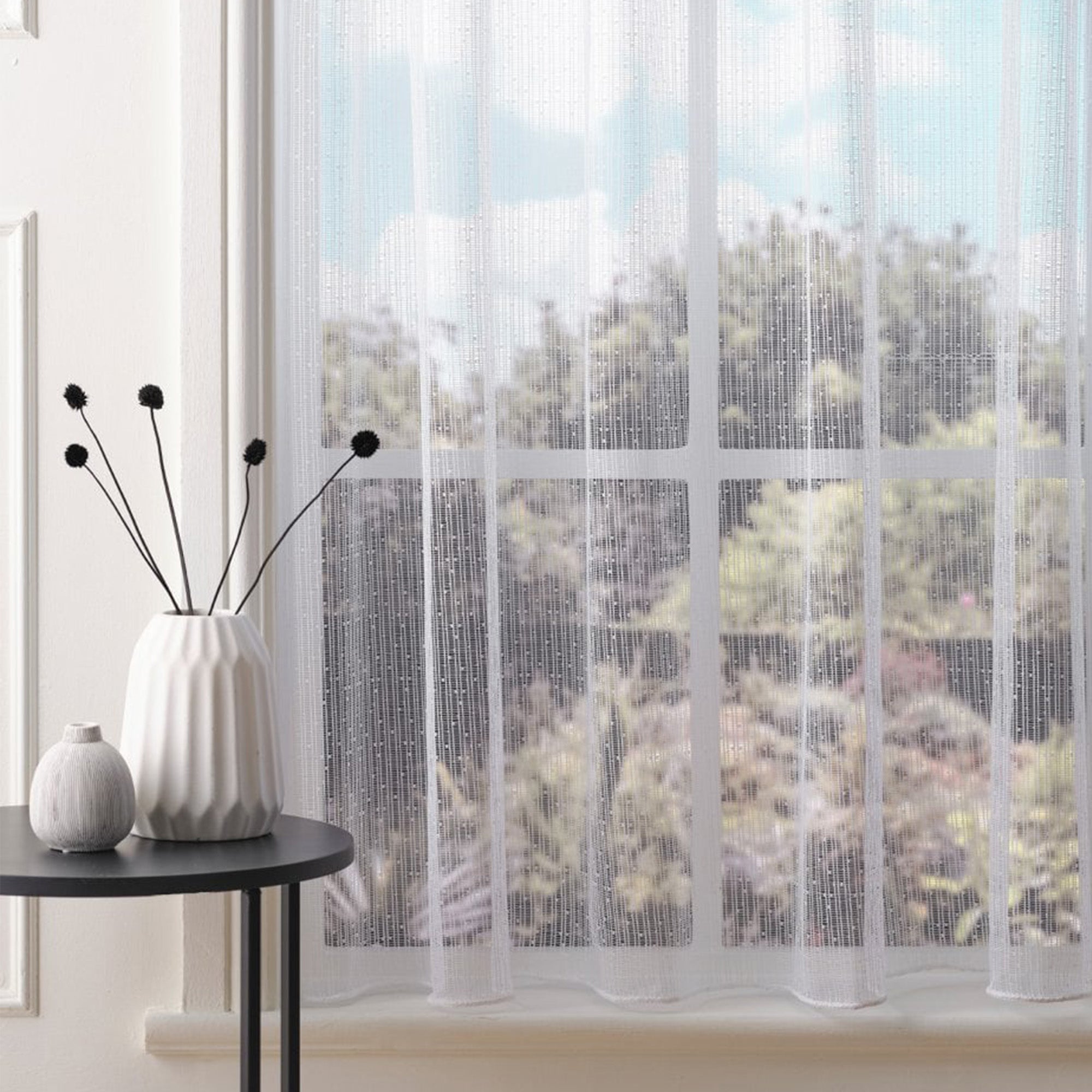White net curtain with vertical subtle stripes and subtle embroidered dots against a blurred outdoor view, placed on a window sill with a table and plant