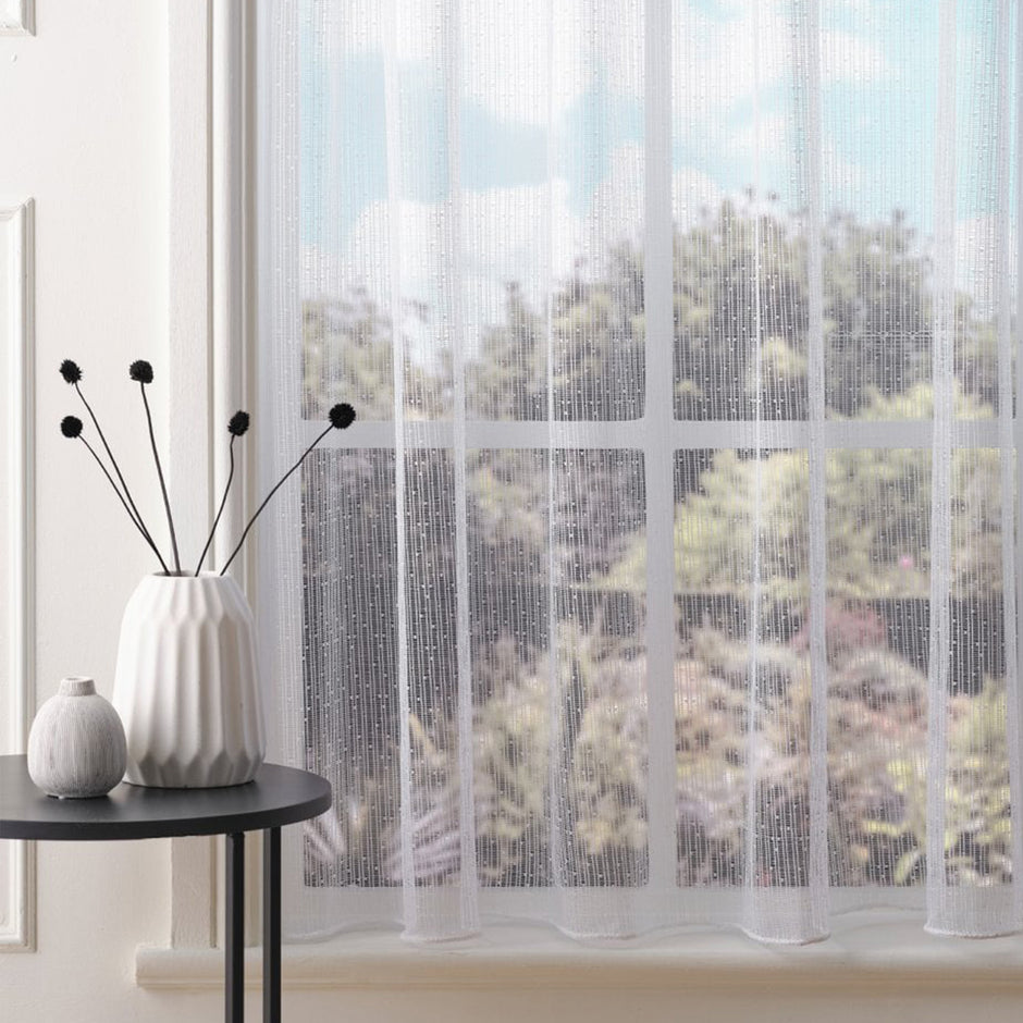 White net curtain with vertical subtle stripes and subtle embroidered dots against a blurred outdoor view, placed on a window sill with a table and plant