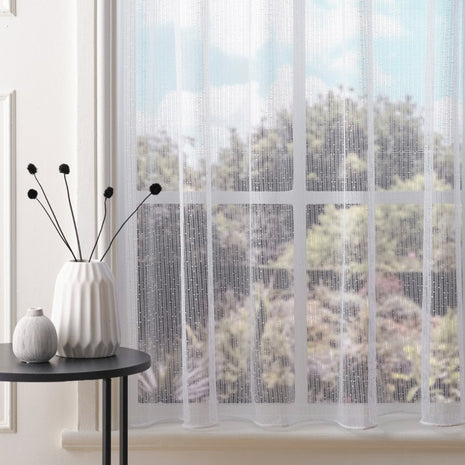 White net curtain with vertical subtle stripes and subtle embroidered dots against a blurred outdoor view, placed on a window sill with a table and plant