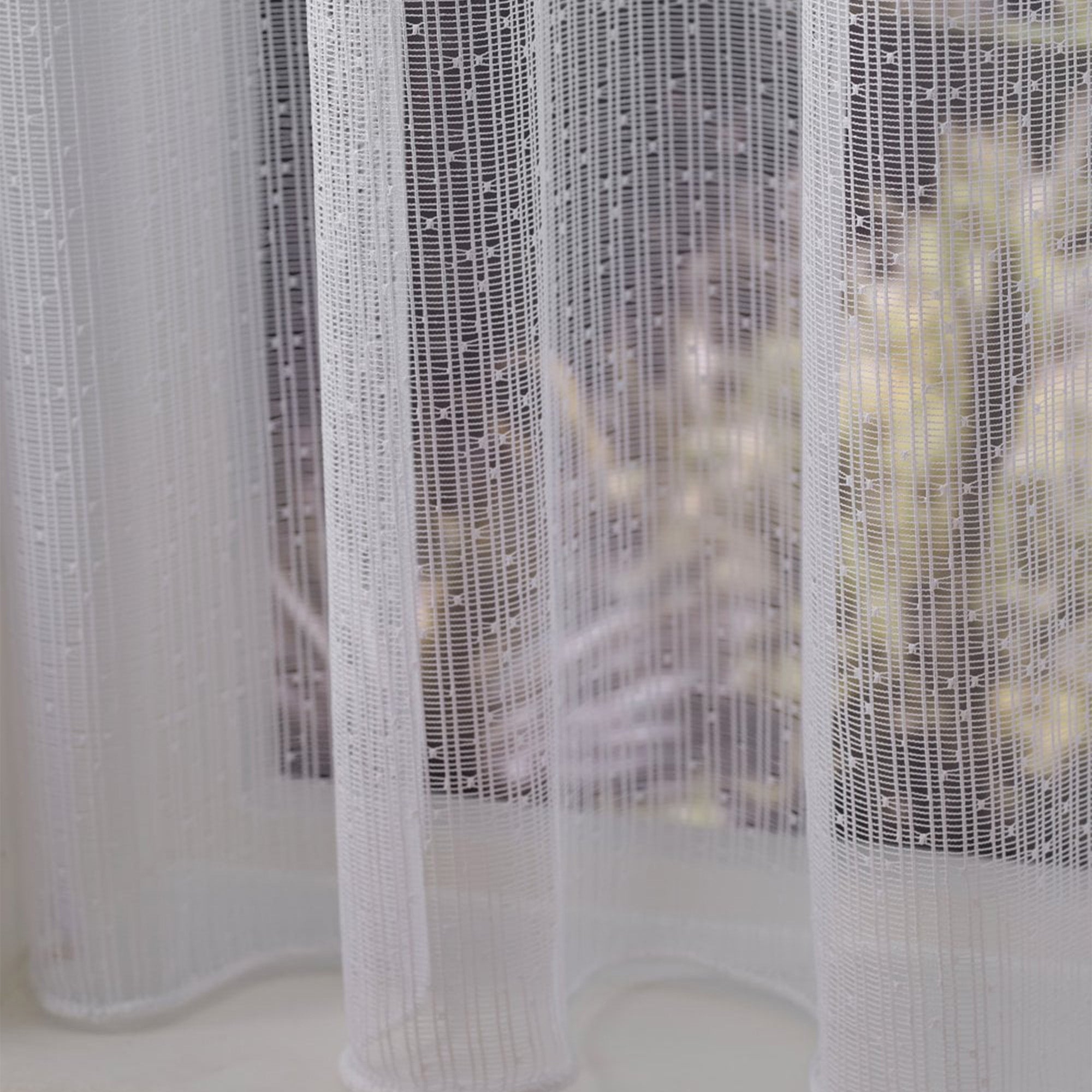 Close-up of a textured white net curtain with vertical sewn lines and dots against a blurred indoor background