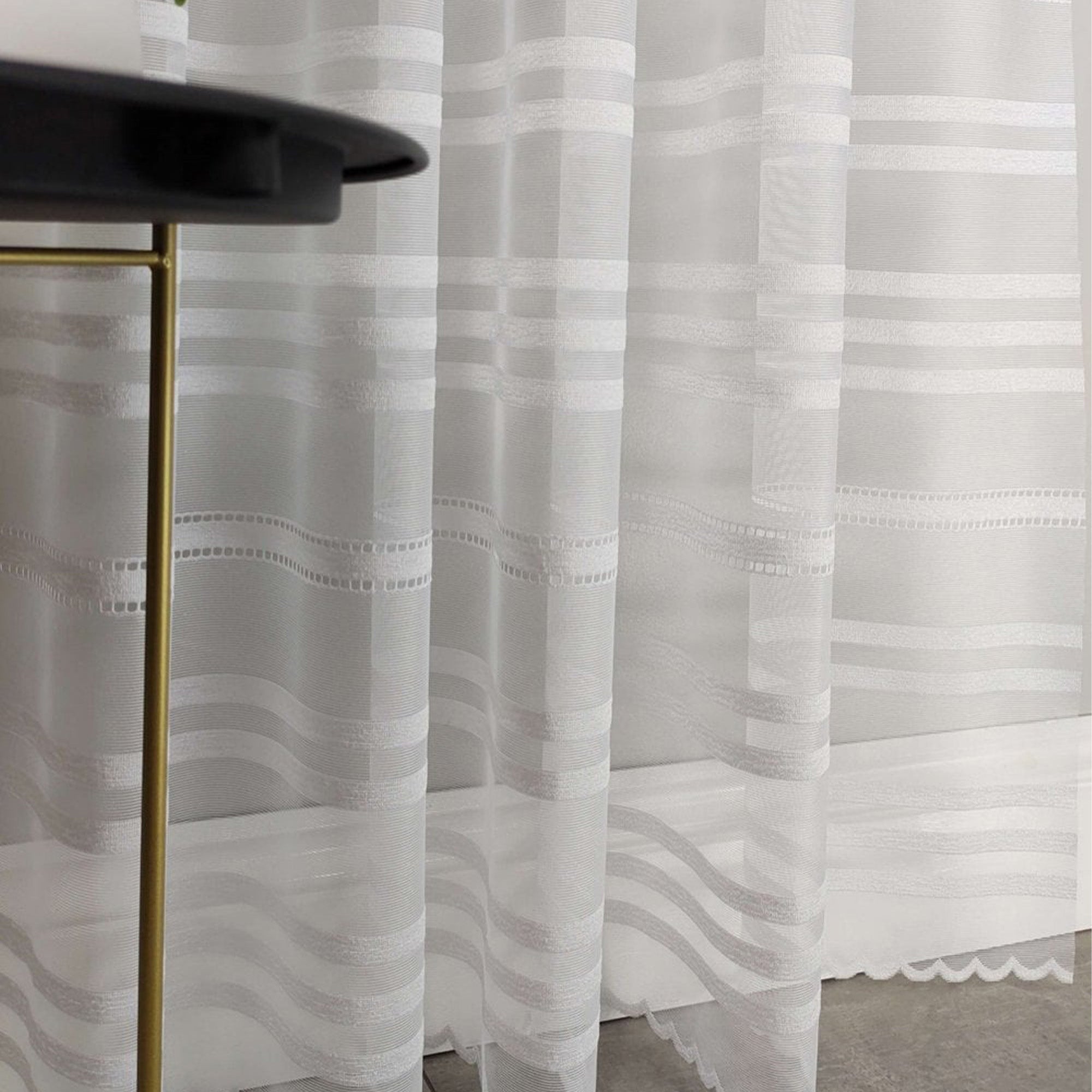 Sheer white curtain with stripe pattern in front of a black table with gold legs.